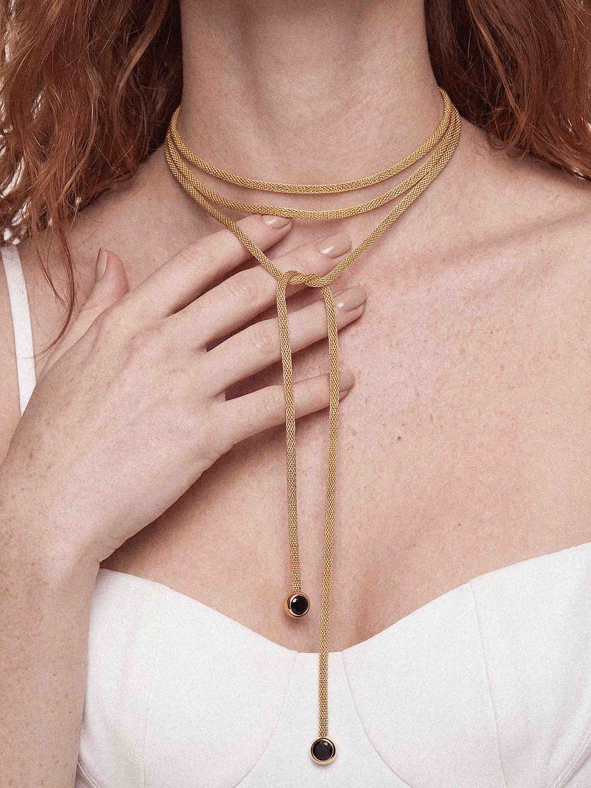 A close-up of a model wearing a multi-layered tan cord necklace with small black circular beads at the ends, paired with a white top.