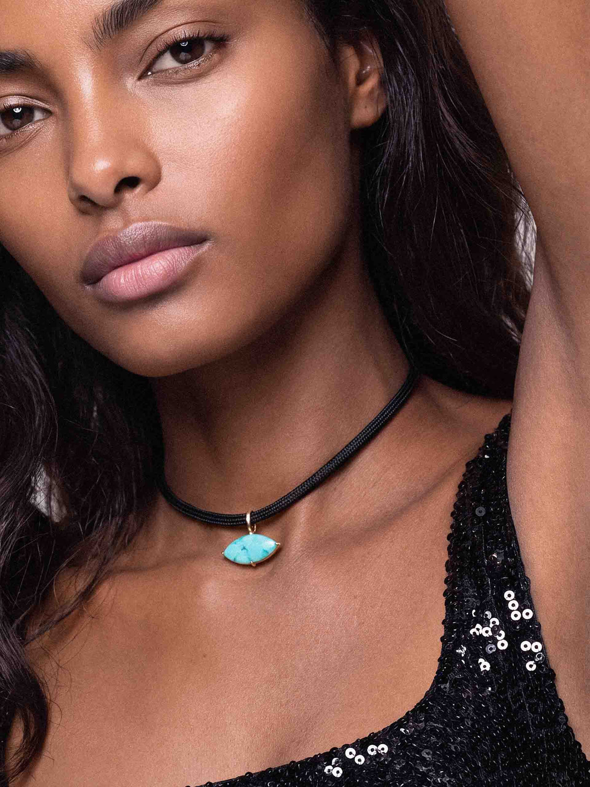 Close-up fashion portrait of a woman wearing a black sequin top and a black cord necklace with a turquoise pendant; her calm gaze feels poised and elegant.
