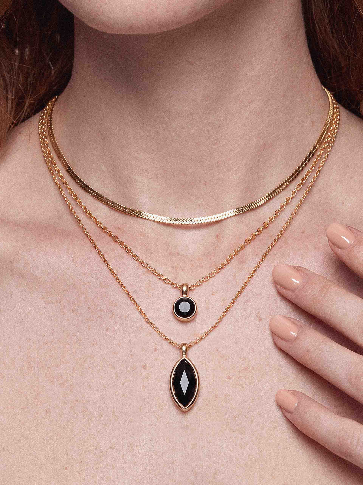 Close-up of a woman wearing layered gold necklaces with two black gemstone pendants, manicured hand on her chest; elegant, refined mood.