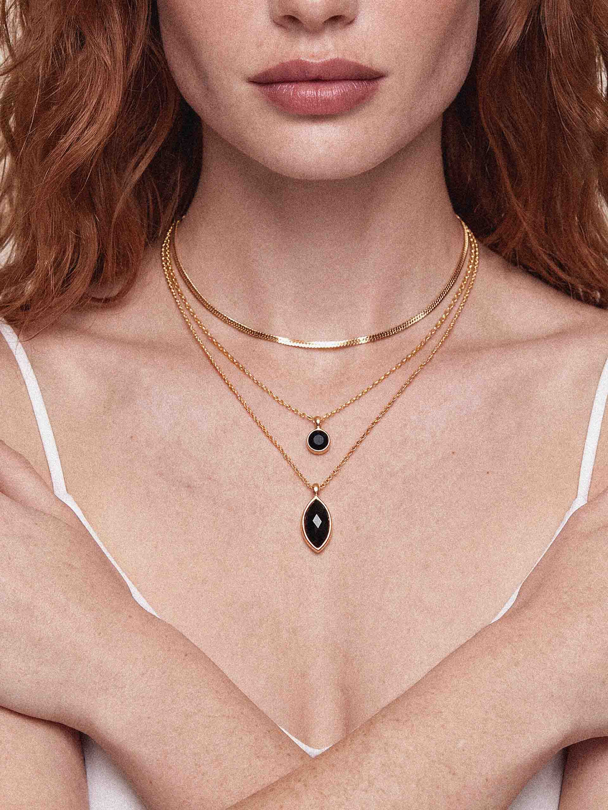 Close-up of a woman with wavy auburn hair wearing layered gold necklaces with black pendants, white straps, and crossed arms.