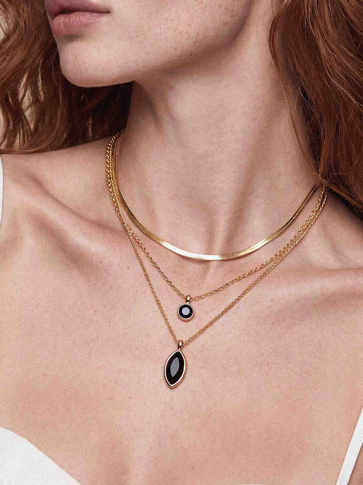 Close-up of a model wearing layered gold necklaces, including a thin chain, a curved band, and two delicate chains with a small round black-and-white pendant and a larger teardrop black pendant.