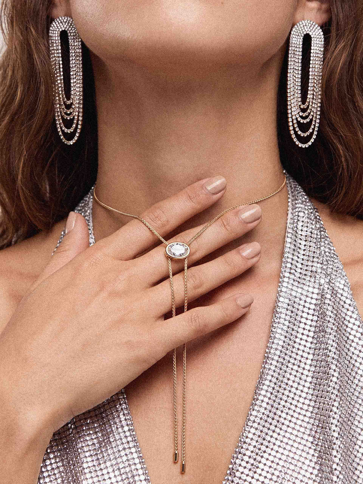 Close-up of a woman wearing long crystal drop earrings, a delicate gold necklace with a gemstone ring pendant, and a shimmering silver outfit; elegant, glamorous styling.
