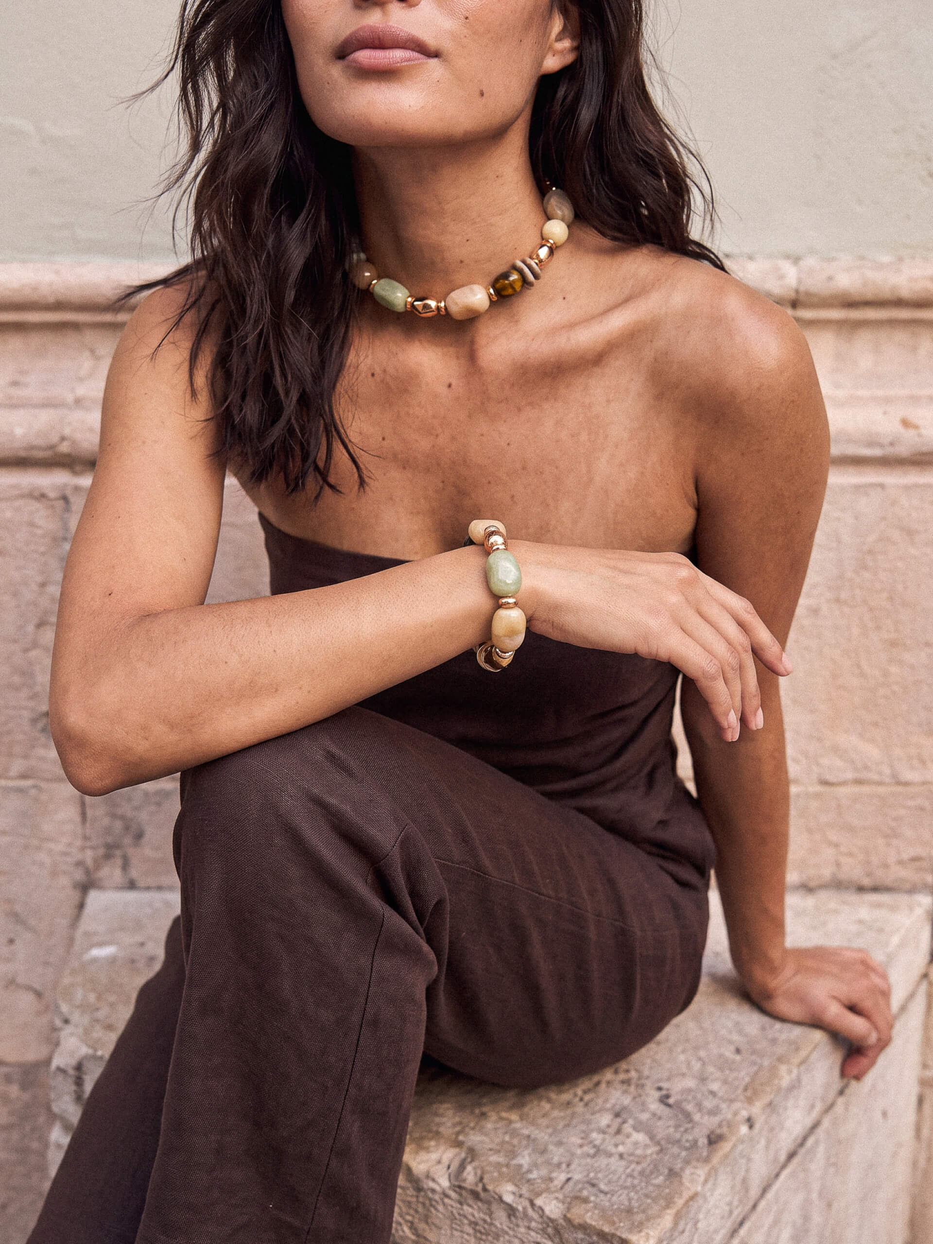 A woman wearing a chunky beaded necklace and matching beaded bracelet, styled with a dark brown strapless top and matching brown pants, sitting on stone steps.