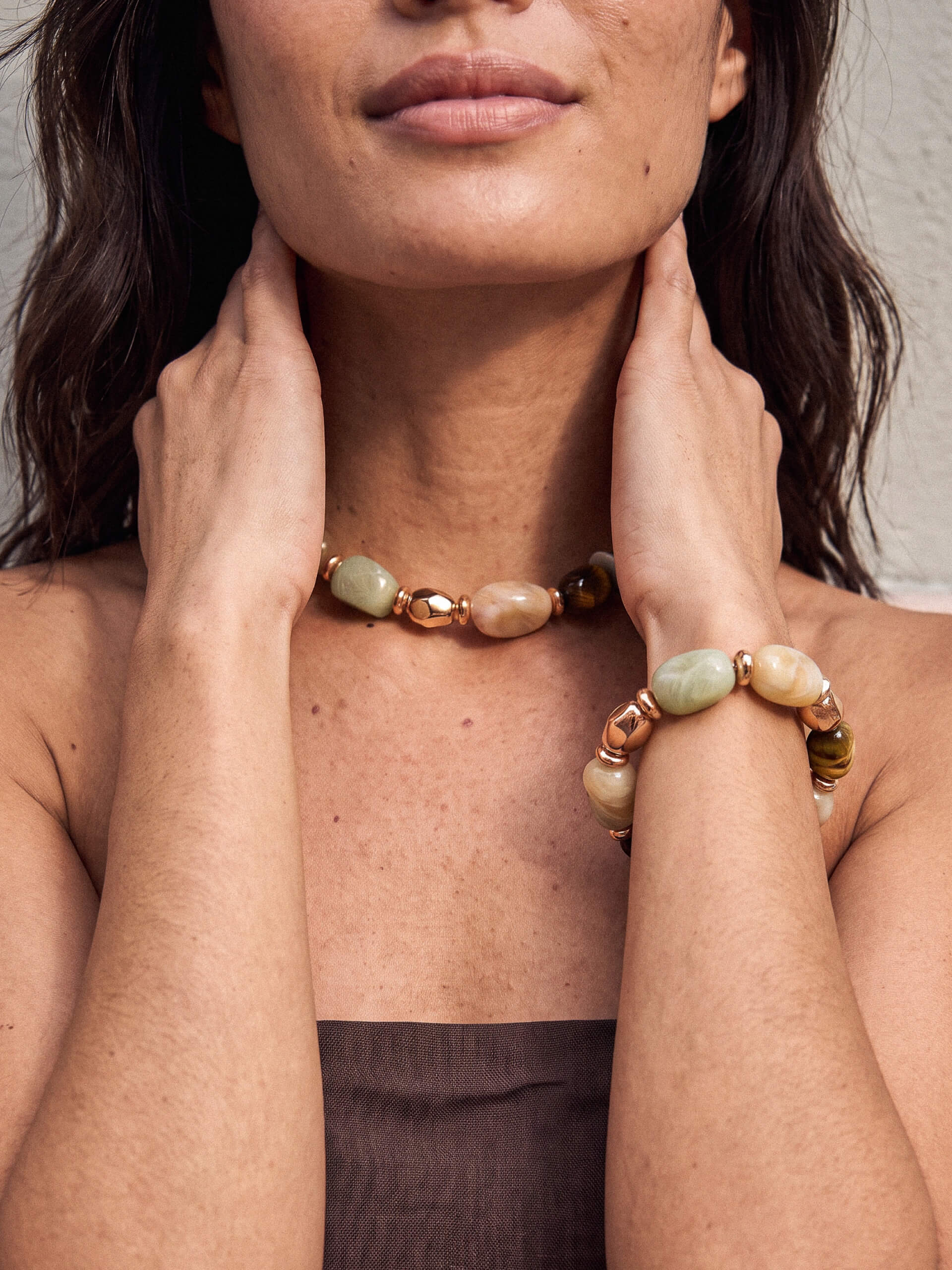 A close-up lifestyle shot of a model wearing a matching beaded necklace and bracelet. The jewelry features polished light green and tan beads with rose gold metallic spacer rings, shown on the model's neck and wrist.
