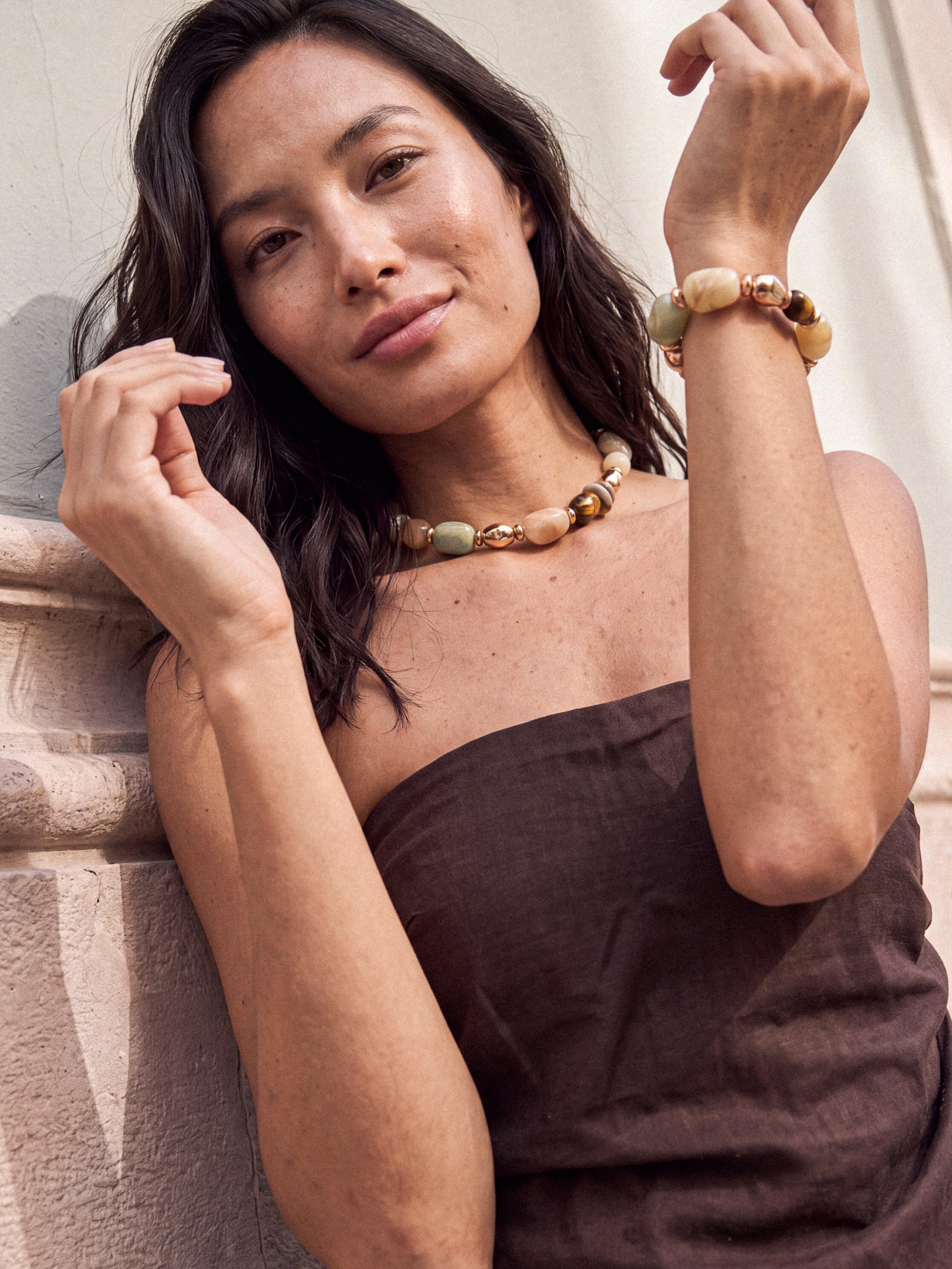 A woman wearing a chunky beaded necklace and matching beaded bracelet, leaning against a stone wall. The jewelry features natural stone beads in earthy tones of beige, brown, and muted green.
