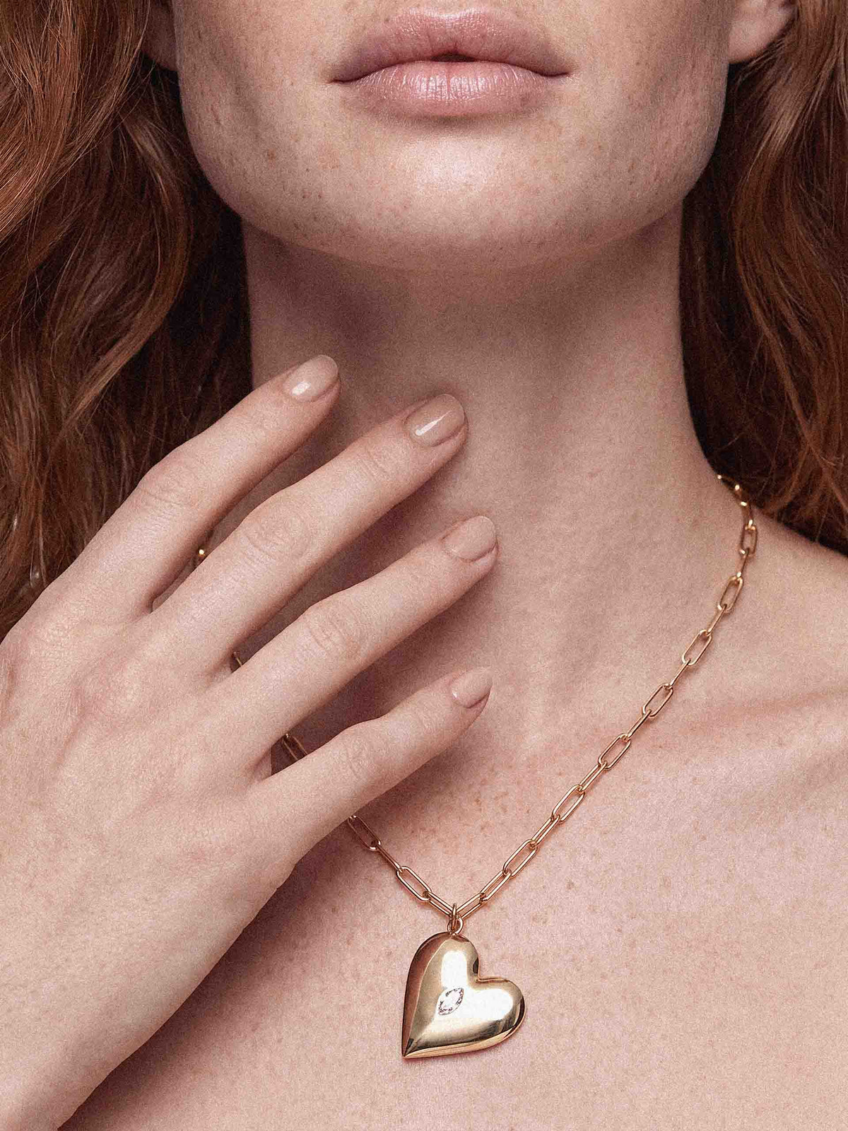 Close-up portrait of a red-haired woman wearing a delicate gold chain with a heart pendant over a white strapless top; warm, joyful mood.