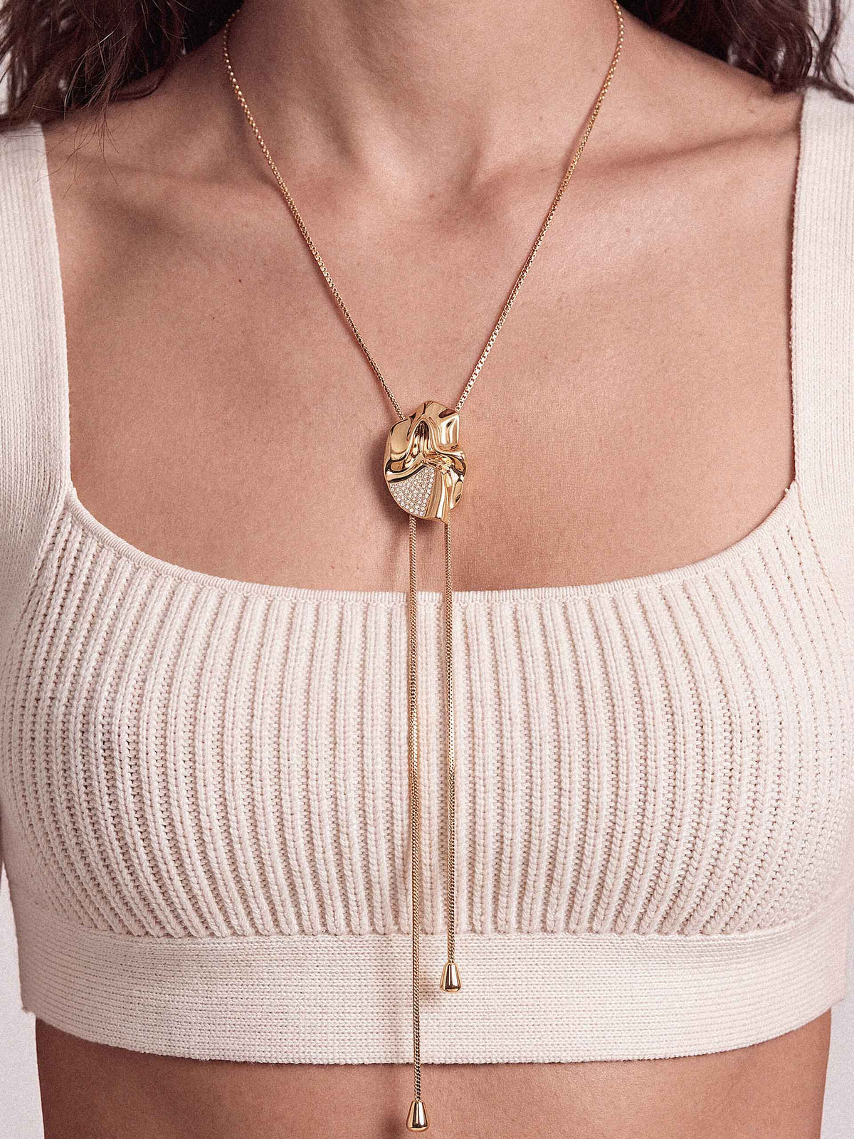 A close-up of a model wearing a gold-toned long necklace with a textured heart-shaped pendant and two long dangling chains, styled over a ribbed cream-colored top.
