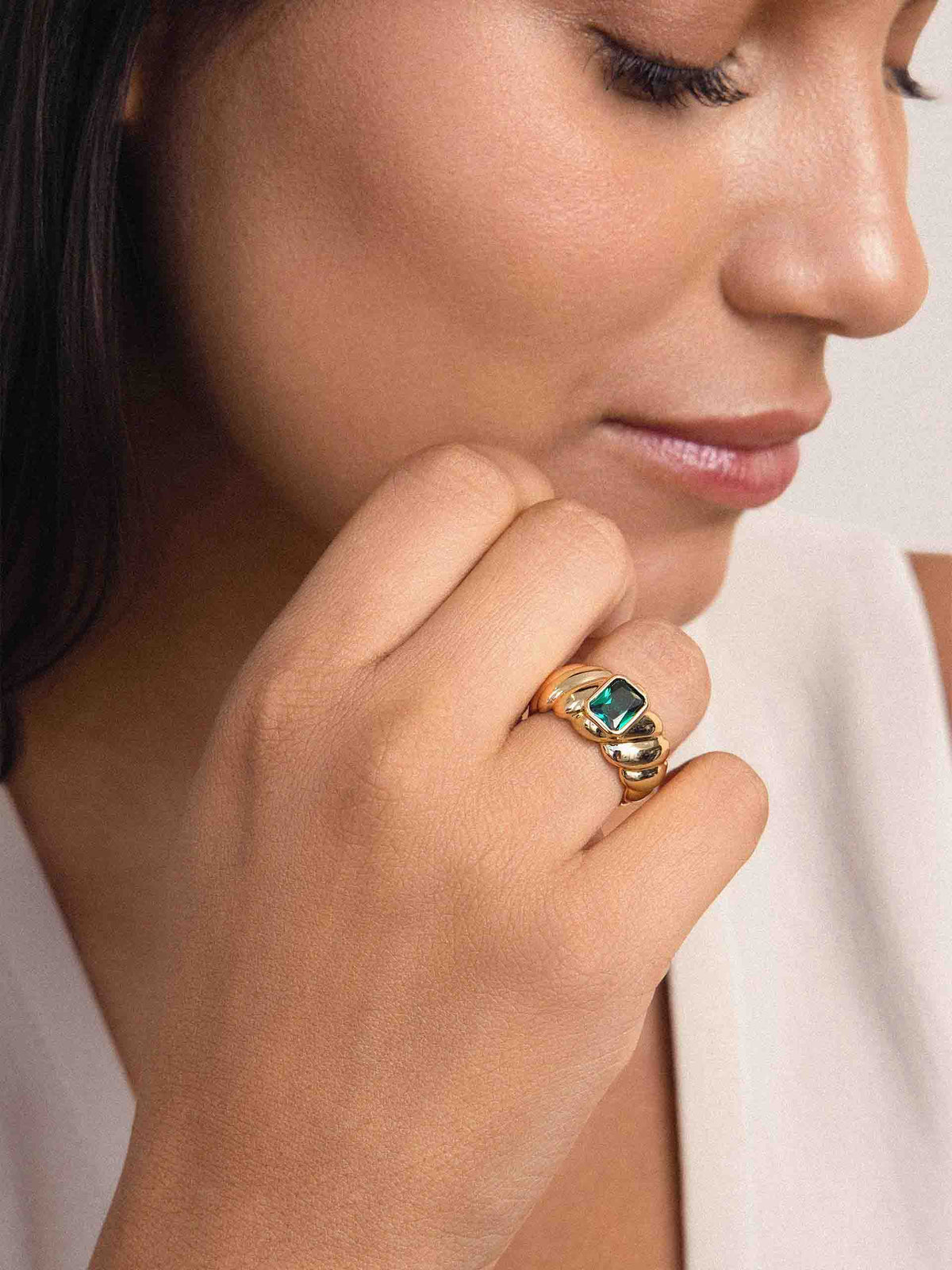 A close-up lifestyle shot of a woman wearing a gold ring with a green rectangular gemstone on her finger, looking down with a soft expression.