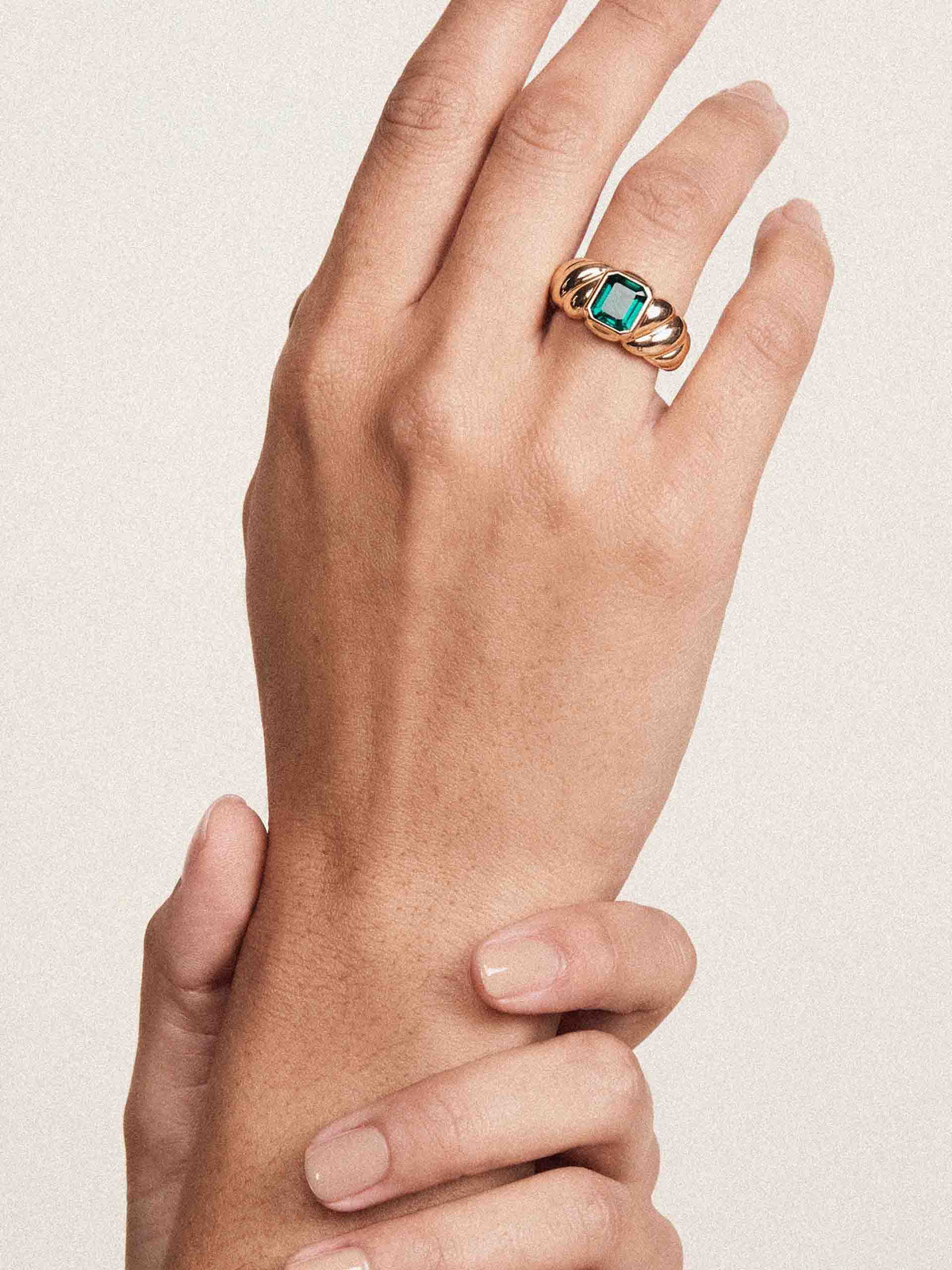A close-up of a hand wearing a gold ring with a square-cut turquoise blue gemstone, set against a plain light beige background.