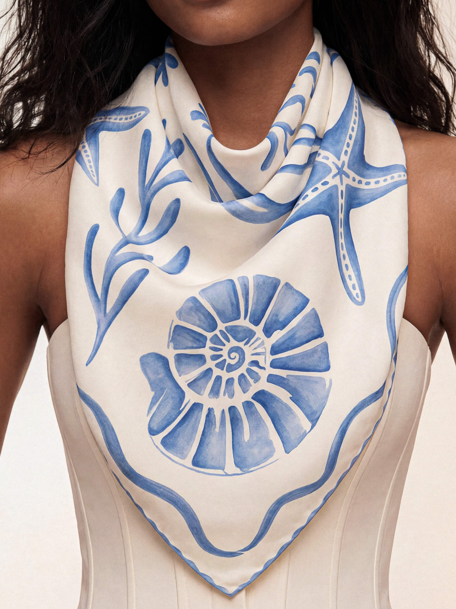 A woman wears a cream scarf with blue seashell and starfish designs over a white strapless top, giving a calm, coastal vibe.
