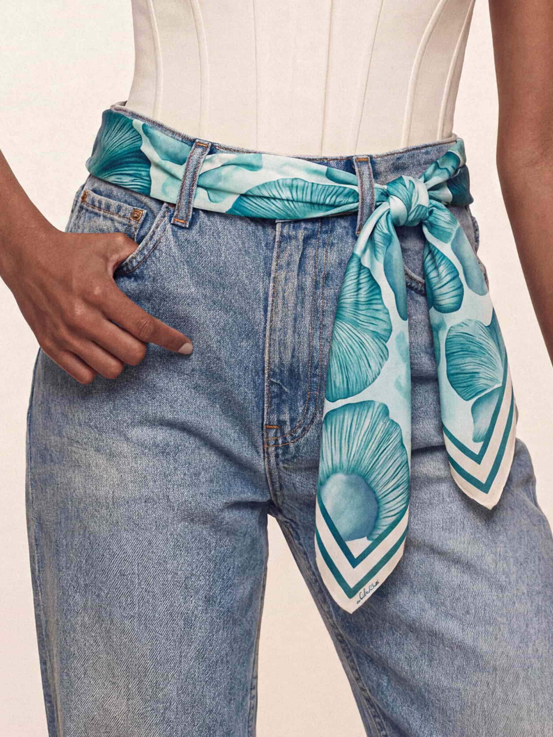 A woman wears a blue floral scarf as a belt. Her hand rests in the pocket, and she wears a white corset top, creating a stylish, casual look.