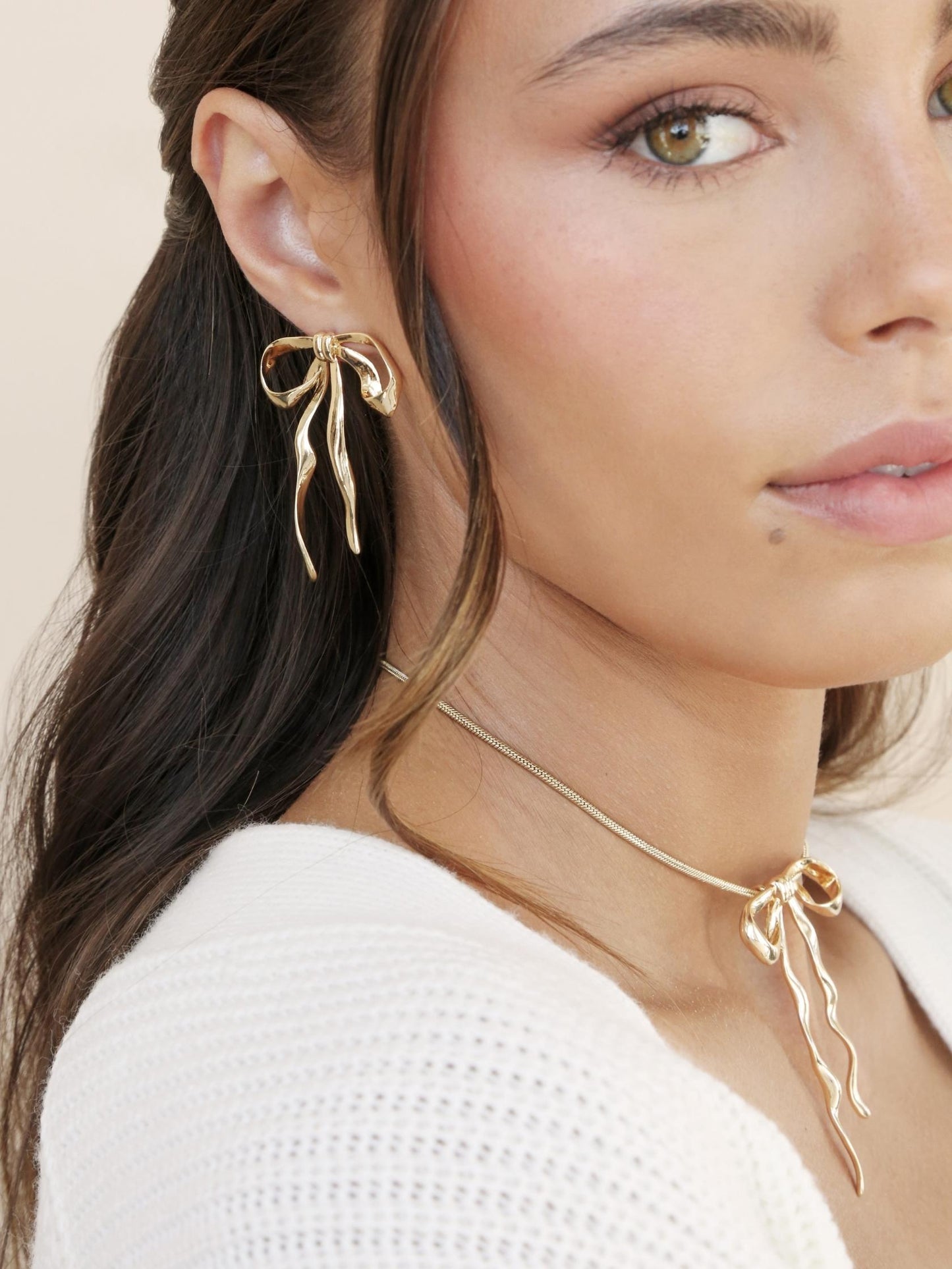 Woman wearing gold bow-shaped earrings and necklace against a beige background