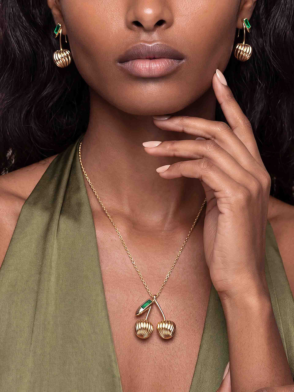 Close-up of a model wearing gold-and-green drop earrings and a matching pendant necklace over an olive dress, posing elegantly with a hand near her face.