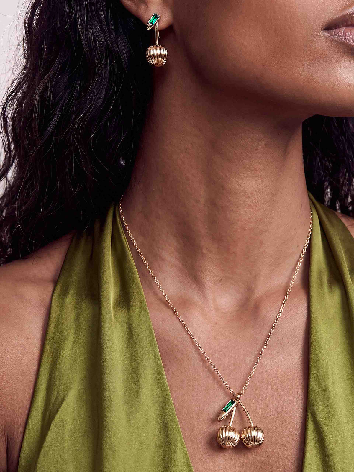 Close-up of a woman wearing a green halter top, gold necklace, and matching dangling earrings with emerald accents and textured spherical charms; elegant, polished look.