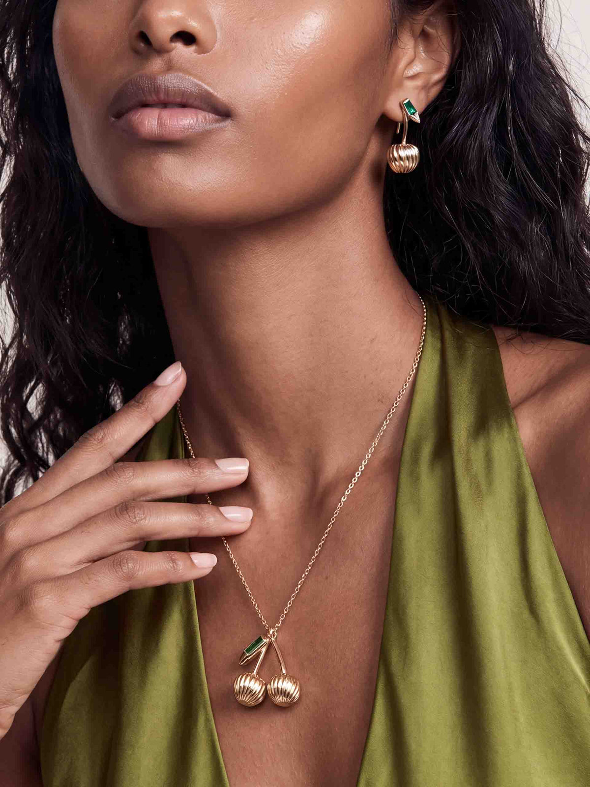 Close-up of a woman in an olive-green satin dress, hand at her neck, displaying gold shell-drop earrings and a matching leaf pendant; elegant, luxe mood.