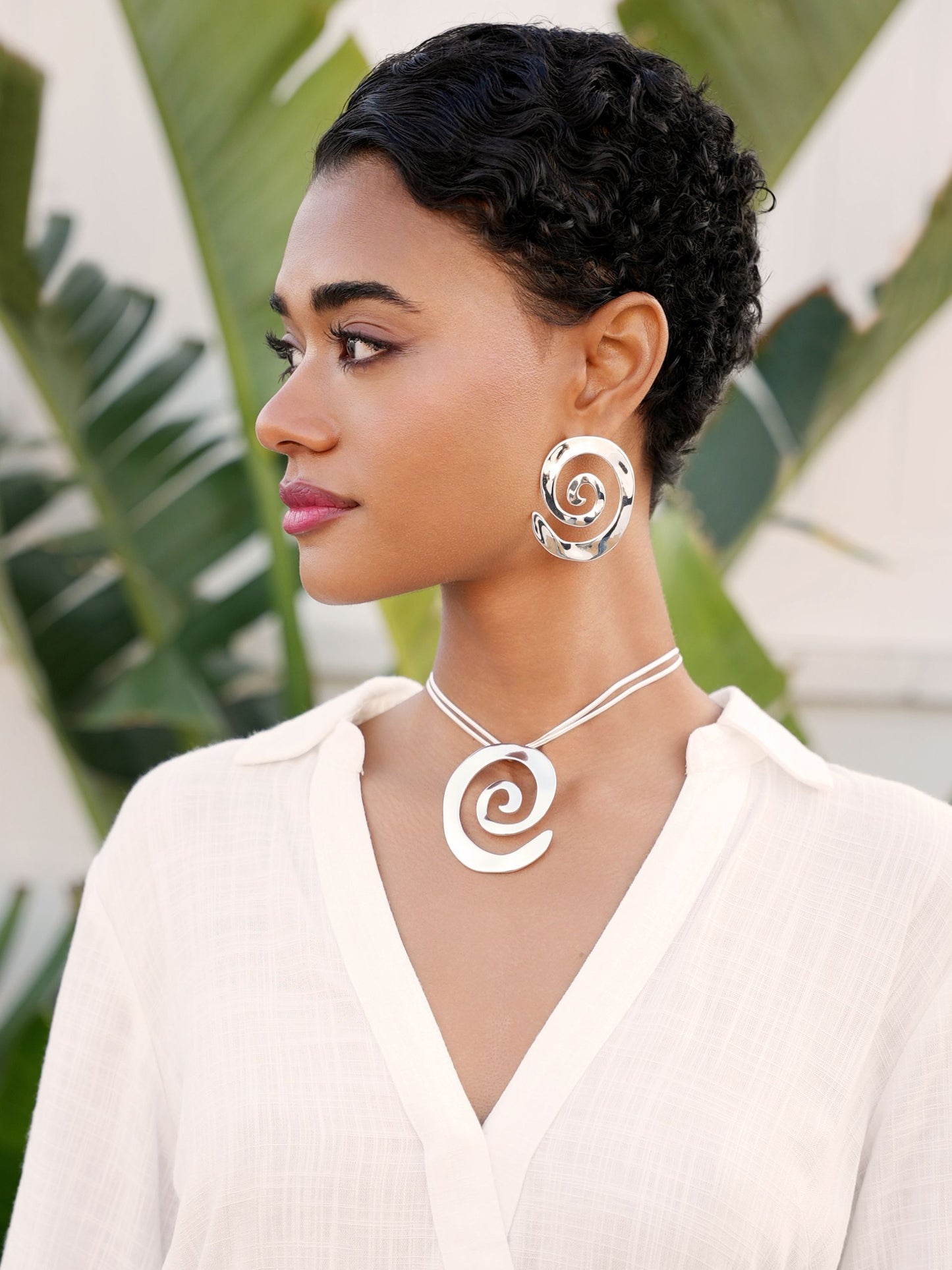 Woman wearing a white top with spiral necklaces and earrings against a blurred green leaf background
