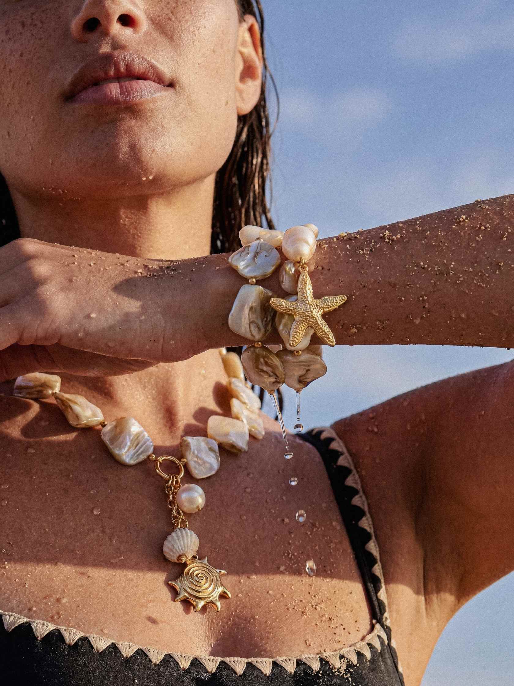 Person wearing a necklace and bracelet with starfish and shell designs against a blue sky.